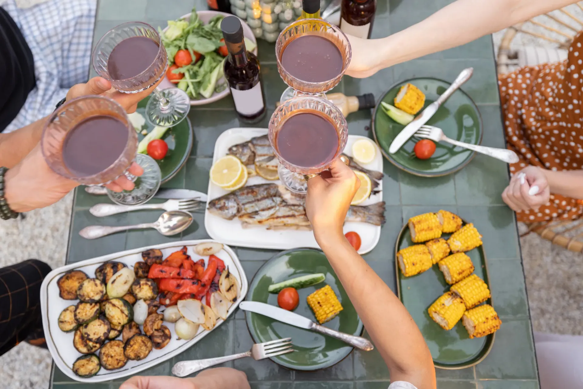 People toasting with red wine at an outdoor table filled with grilled vegetables and fish.