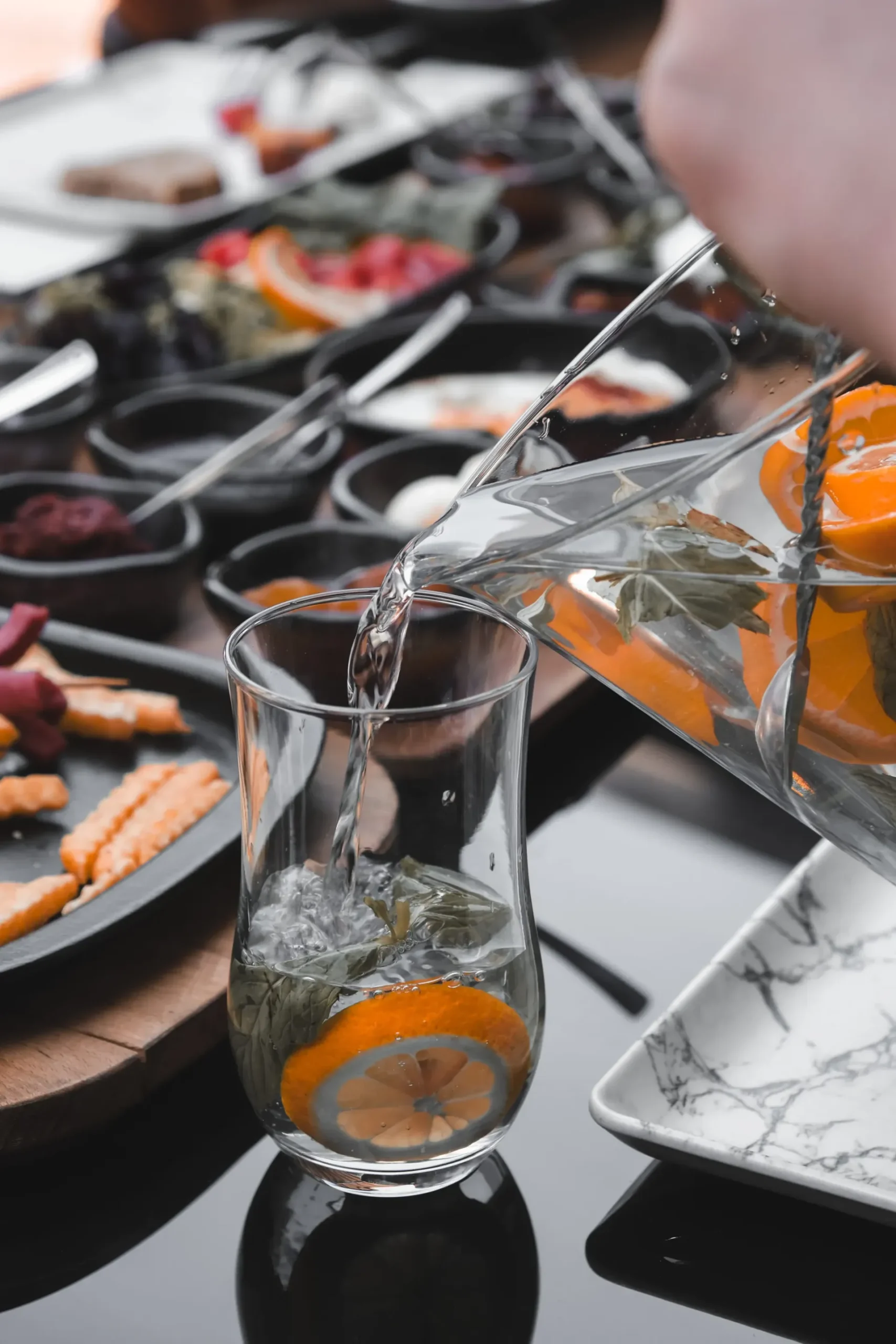 Person pouring water into glass with citrus and herbs on table with food trays.