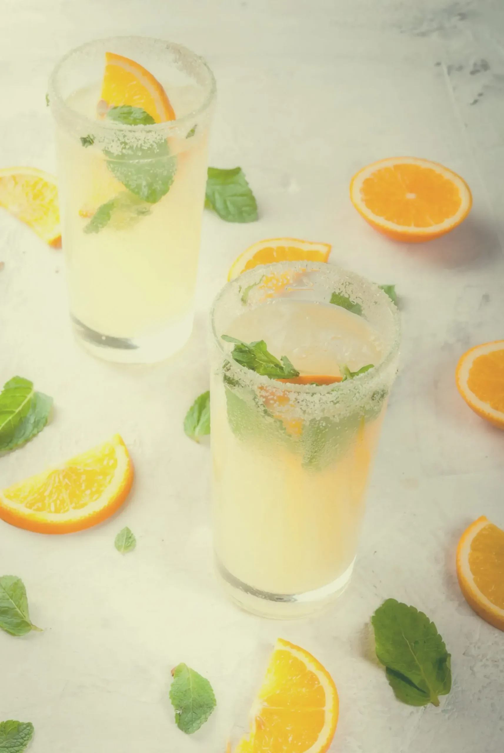 Two glasses of citrusy drinks with mint leaves and orange slices on a white surface.