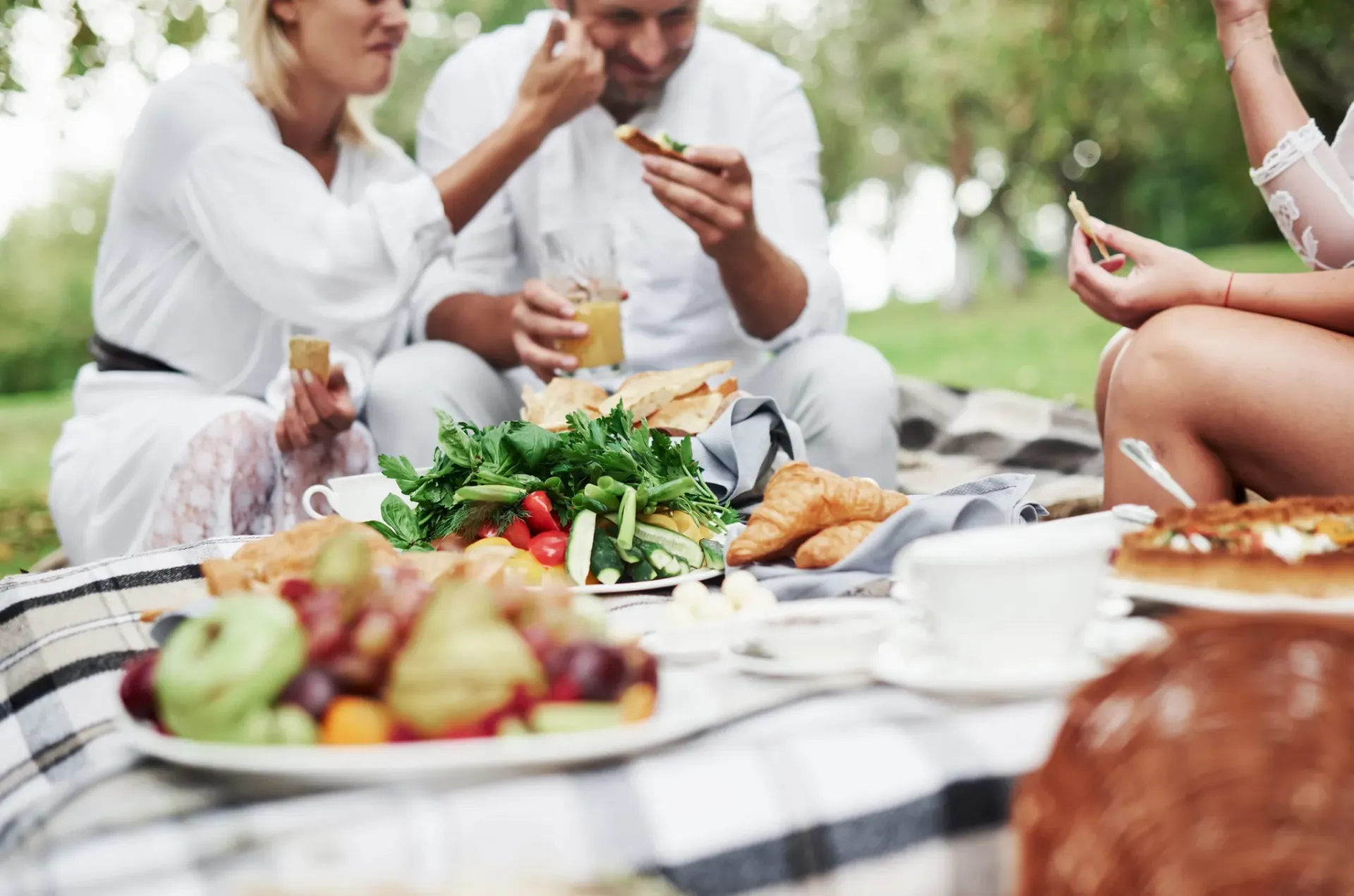 Three people enjoying a picnic with fruits, salads, and bread on a blanket in a park.