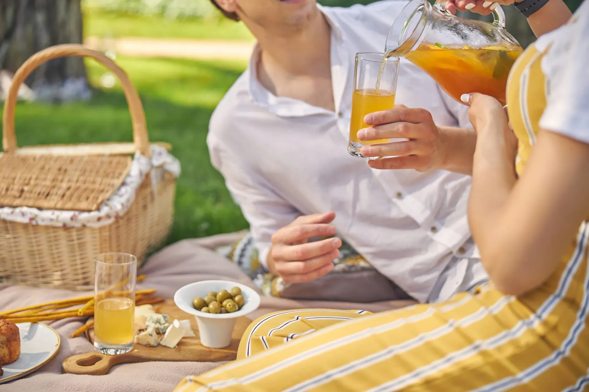A couple enjoys a picnic with drinks and snacks on a blanket in a park.