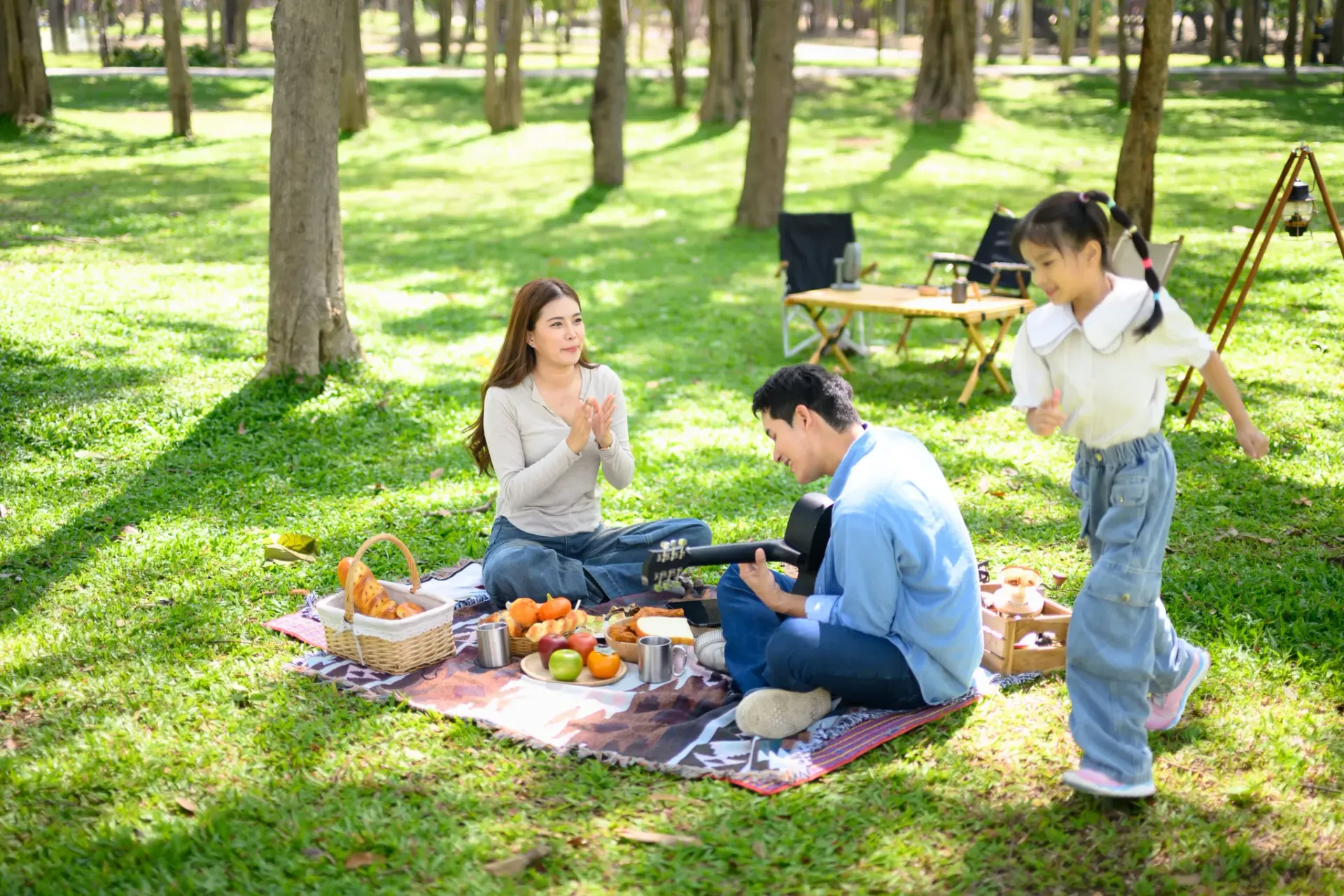 A family enjoys a picnic in a sunny park with a blanket, food, and camping chairs.