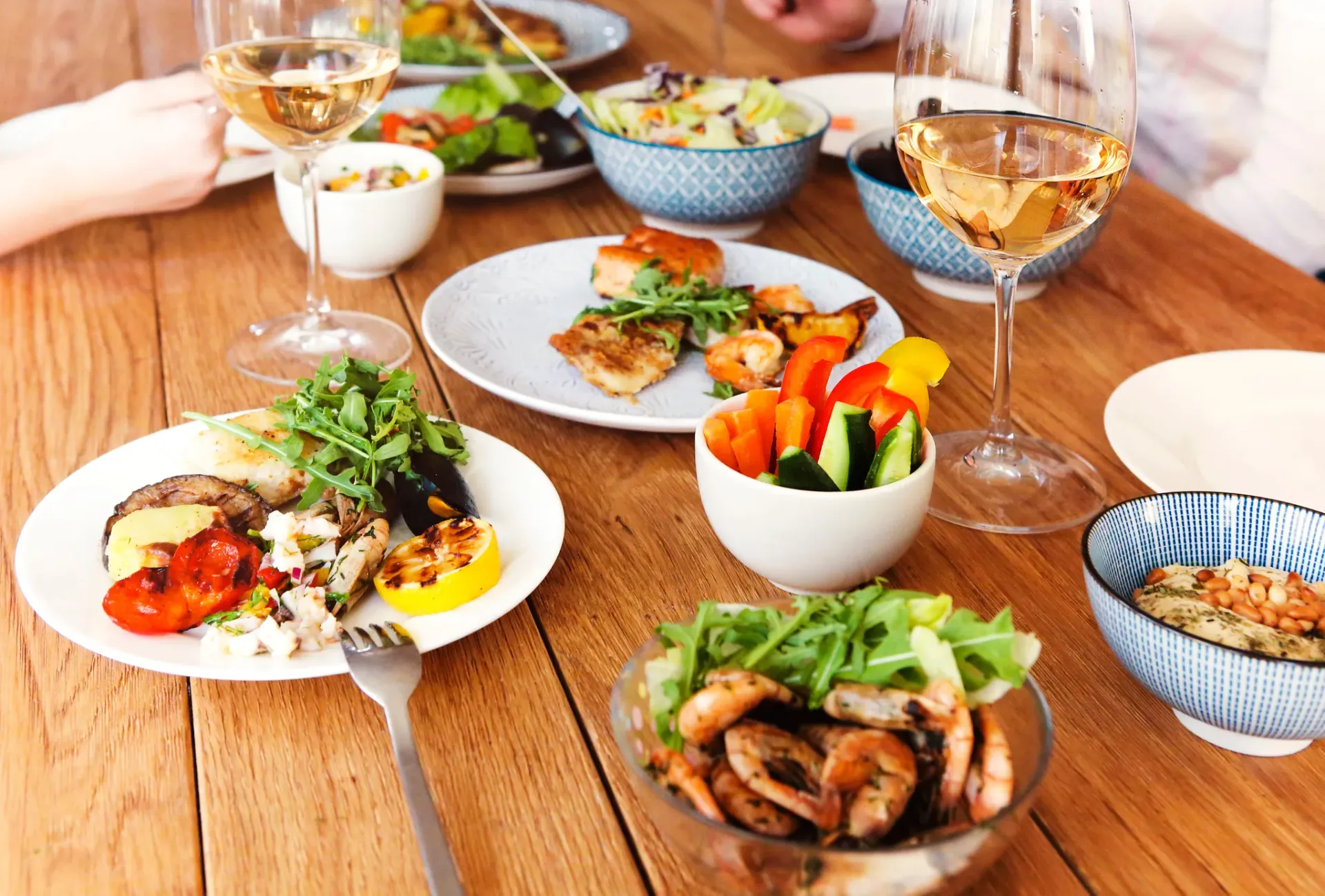 A wooden table set for two with plates of grilled shrimp, vegetables, and white wine glasses.