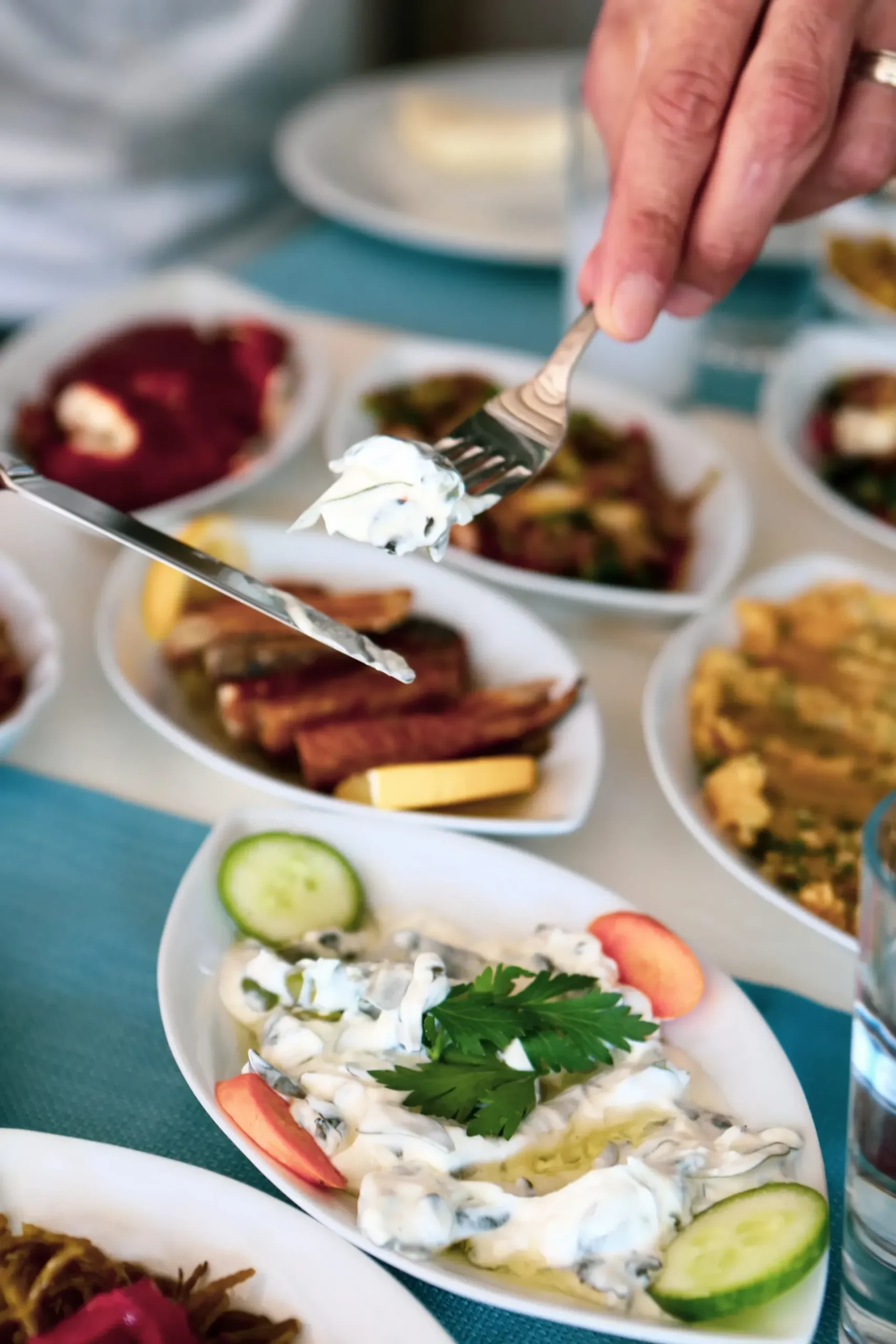 A hand holds a fork with yogurt over plates of various dishes including fish and vegetables on a table.